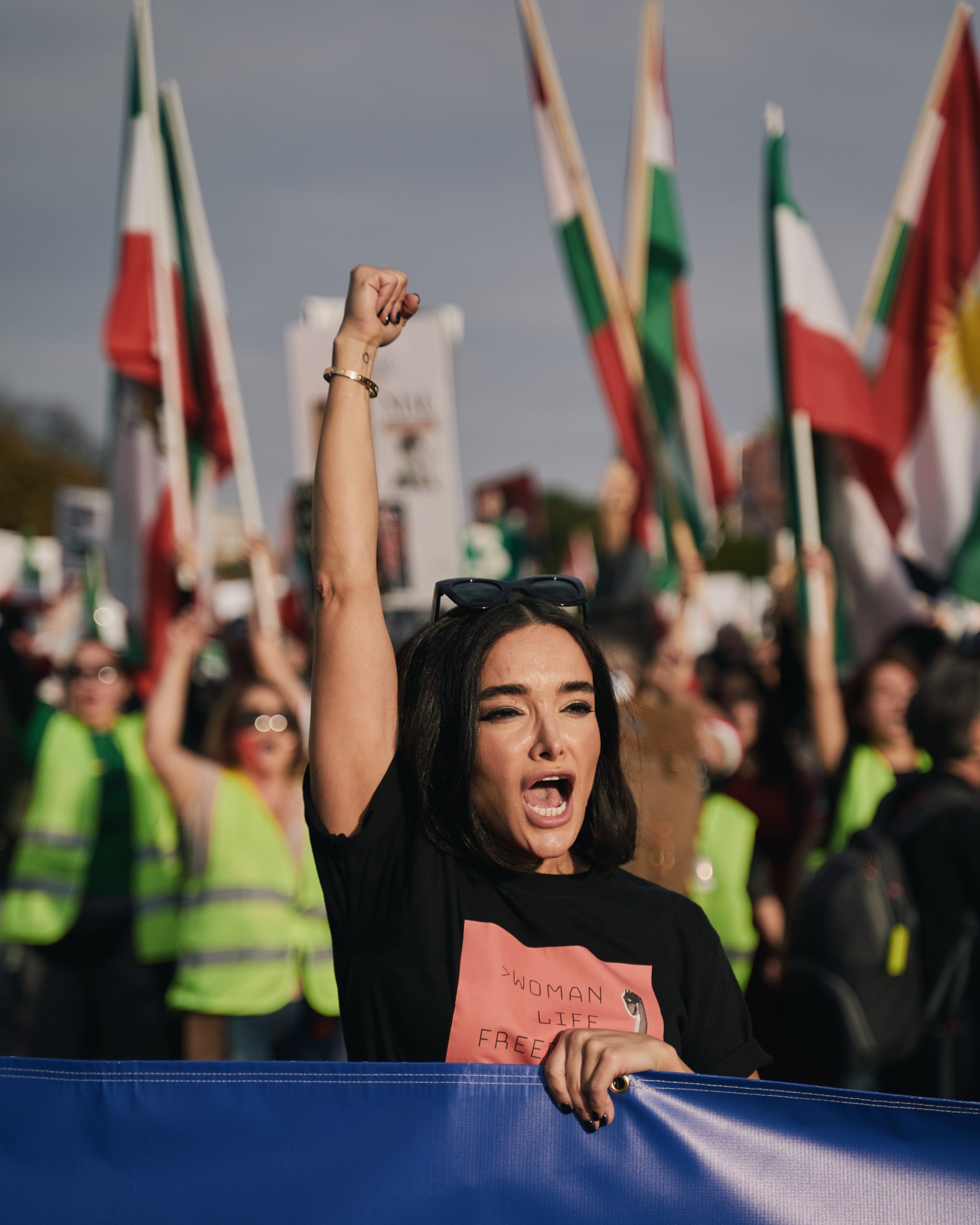 A woman with a "woman, life, freedom" t-shirt has her fist in the air at a protest. There are people with high-vis jackets and Iranian flags behind her.